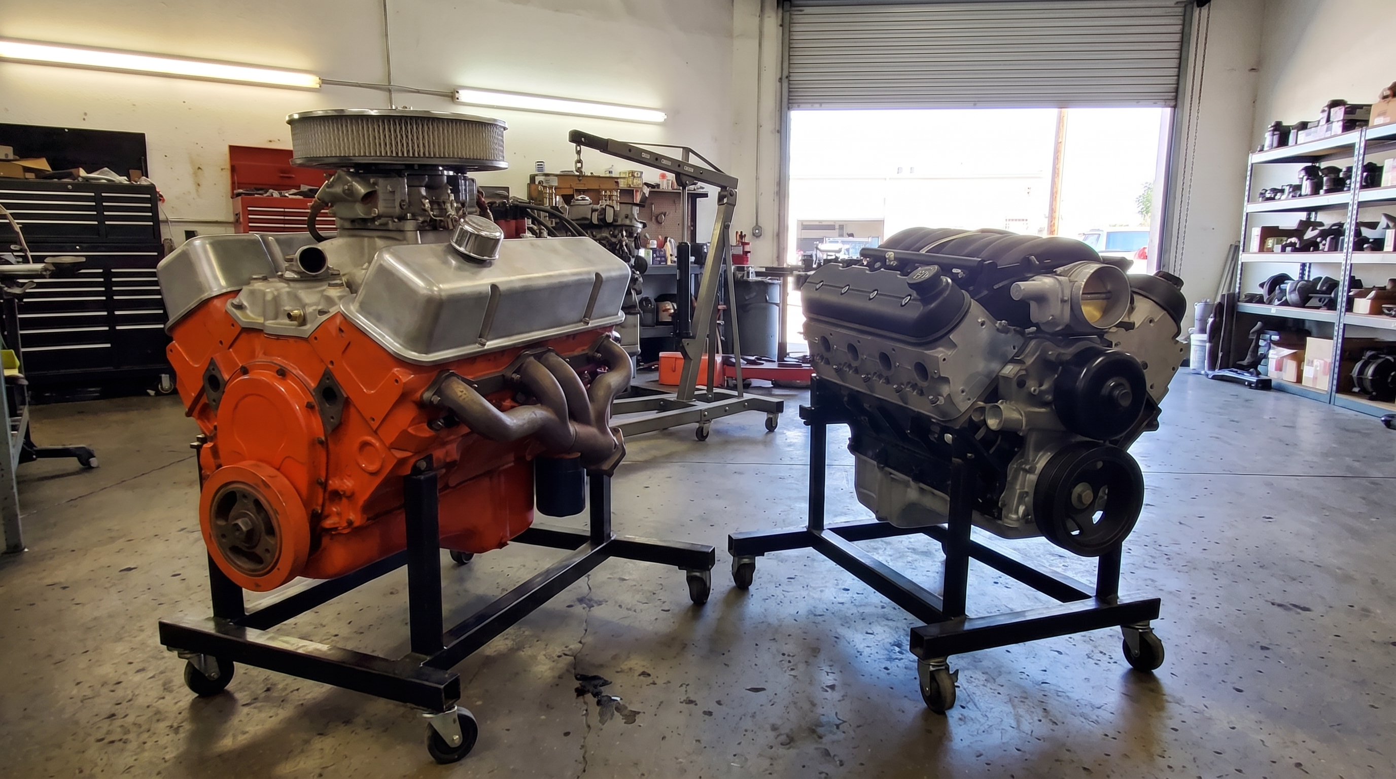 A cast iron Chevrolet big block V8 painted Chevrolet engine orange sits on an engine stand beside a modern LS style V8, showing the size difference between the two engines