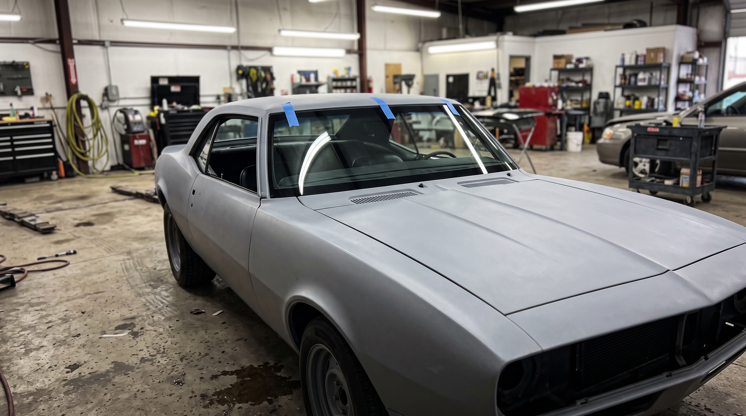 Classic muscle car in primer inside a restoration shop with blue painter tape around a freshly set flush-mount windshield