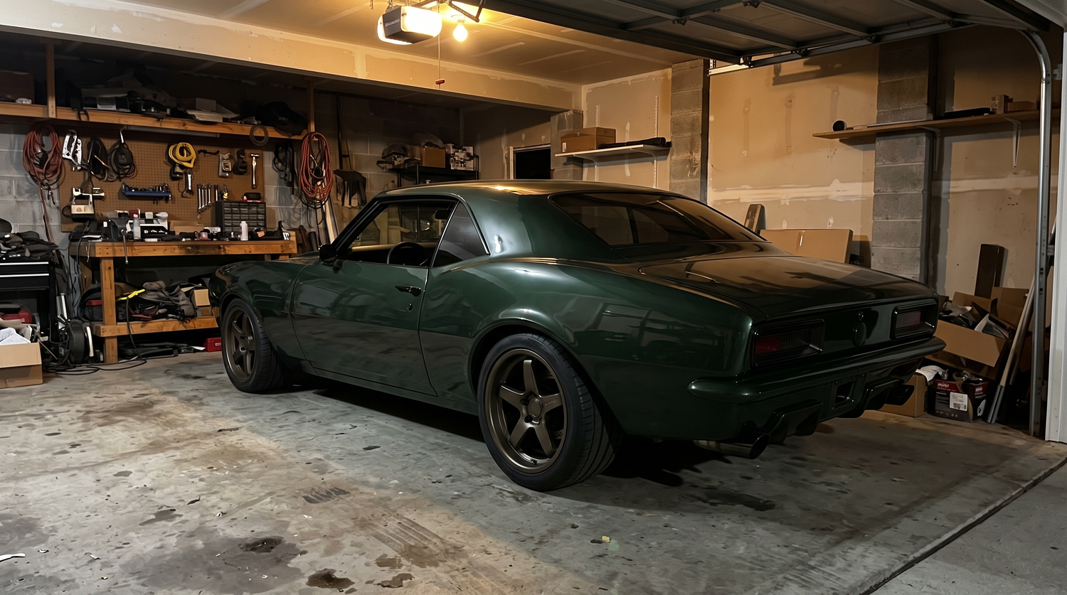 Dark green pro-touring muscle coupe parked at night in a home garage under warm light with flush-mount glass